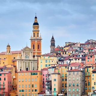 Colourful hillside town with pastel buildings and a prominent church tower rising above the rooftops under a cloudy sky.