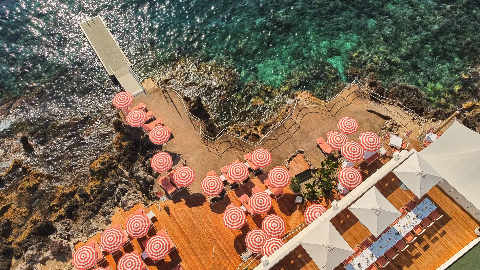 Vue aérienne d’une terrasse en bord de mer avec des parasols rayés rouge et blanc et des transats, au bord d’une eau turquoise et d’un rivage rocheux.