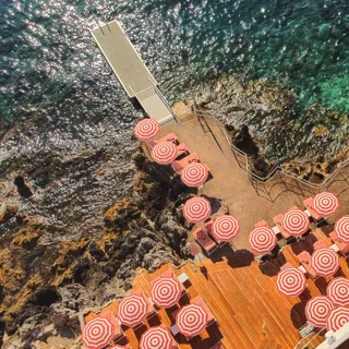 Vue aérienne d’une terrasse en bord de mer avec des parasols rayés rouge et blanc et des transats, au bord d’une eau turquoise et d’un rivage rocheux.