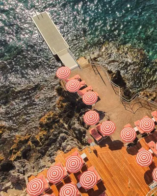 Vue aérienne d’une terrasse en bord de mer avec des parasols rayés rouge et blanc et des transats, au bord d’une eau turquoise et d’un rivage rocheux.