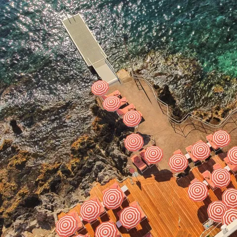 Vue aérienne d’une terrasse en bord de mer avec des parasols rayés rouge et blanc et des transats, au bord d’une eau turquoise et d’un rivage rocheux.