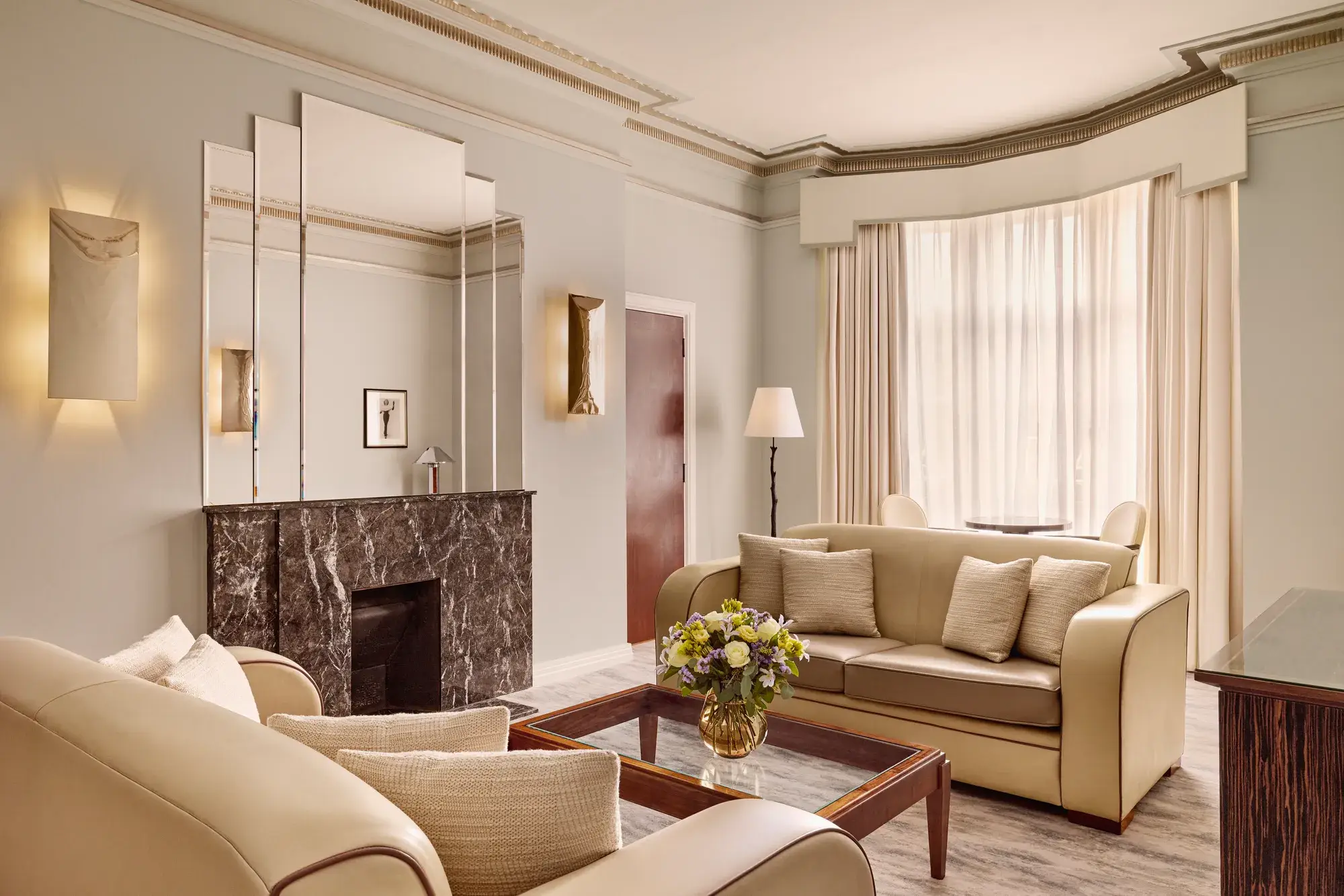 Contemporary living room with cream leather sofas, a dark marble fireplace with mirrored panels above, and a glass coffee table with a floral arrangement, softly lit by natural light through sheer curtains.