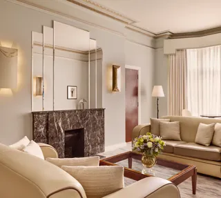 Contemporary living room with cream leather sofas, a dark marble fireplace with mirrored panels above, and a glass coffee table with a floral arrangement, softly lit by natural light through sheer curtains.