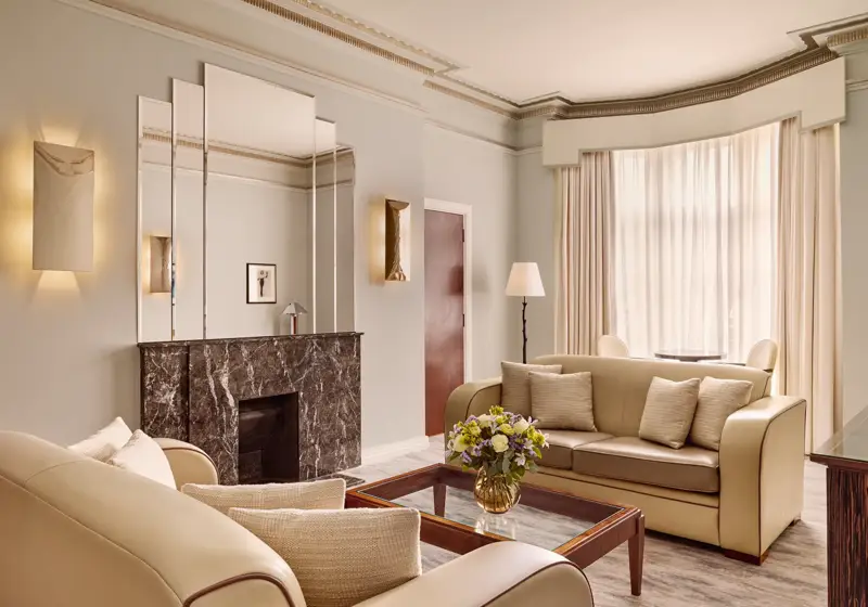 Contemporary living room with cream leather sofas, a dark marble fireplace with mirrored panels above, and a glass coffee table with a floral arrangement, softly lit by natural light through sheer curtains.