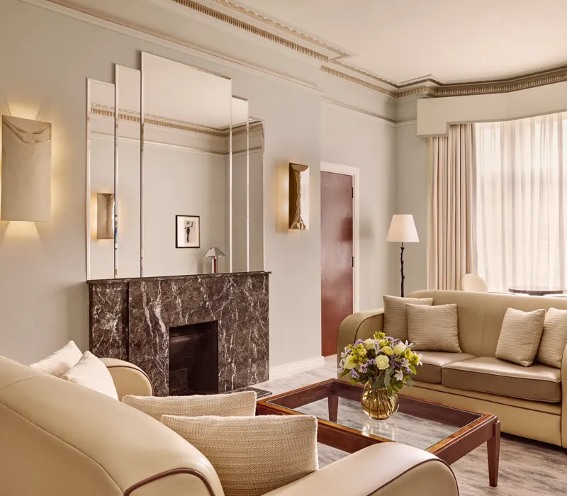 Contemporary living room with cream leather sofas, a dark marble fireplace with mirrored panels above, and a glass coffee table with a floral arrangement, softly lit by natural light through sheer curtains.
