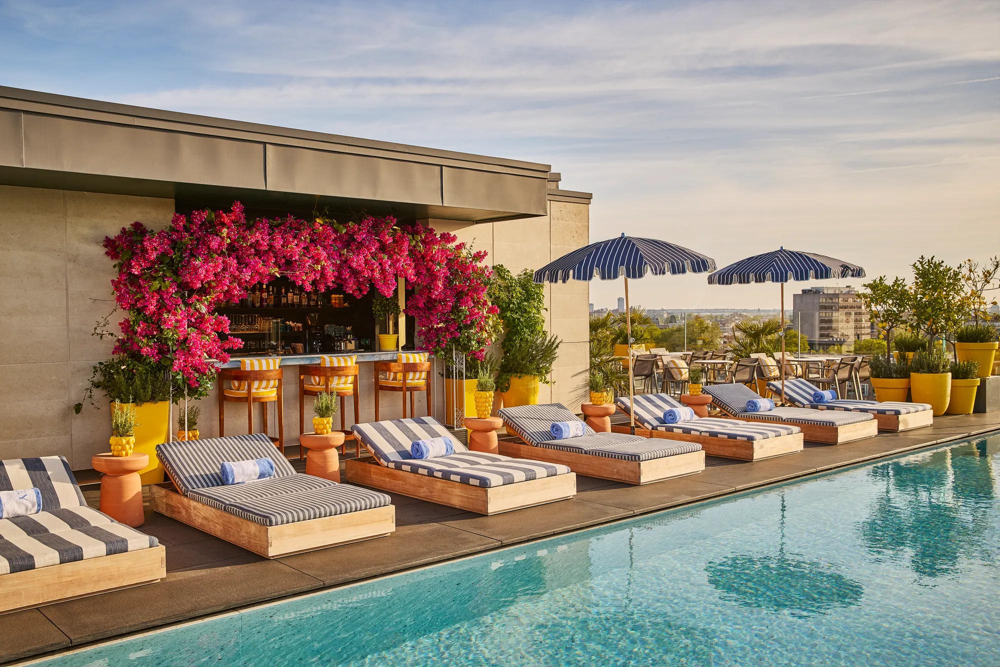 Rooftop pool with striped loungers, blue parasols and a flower-covered bar overlooking the city skyline.