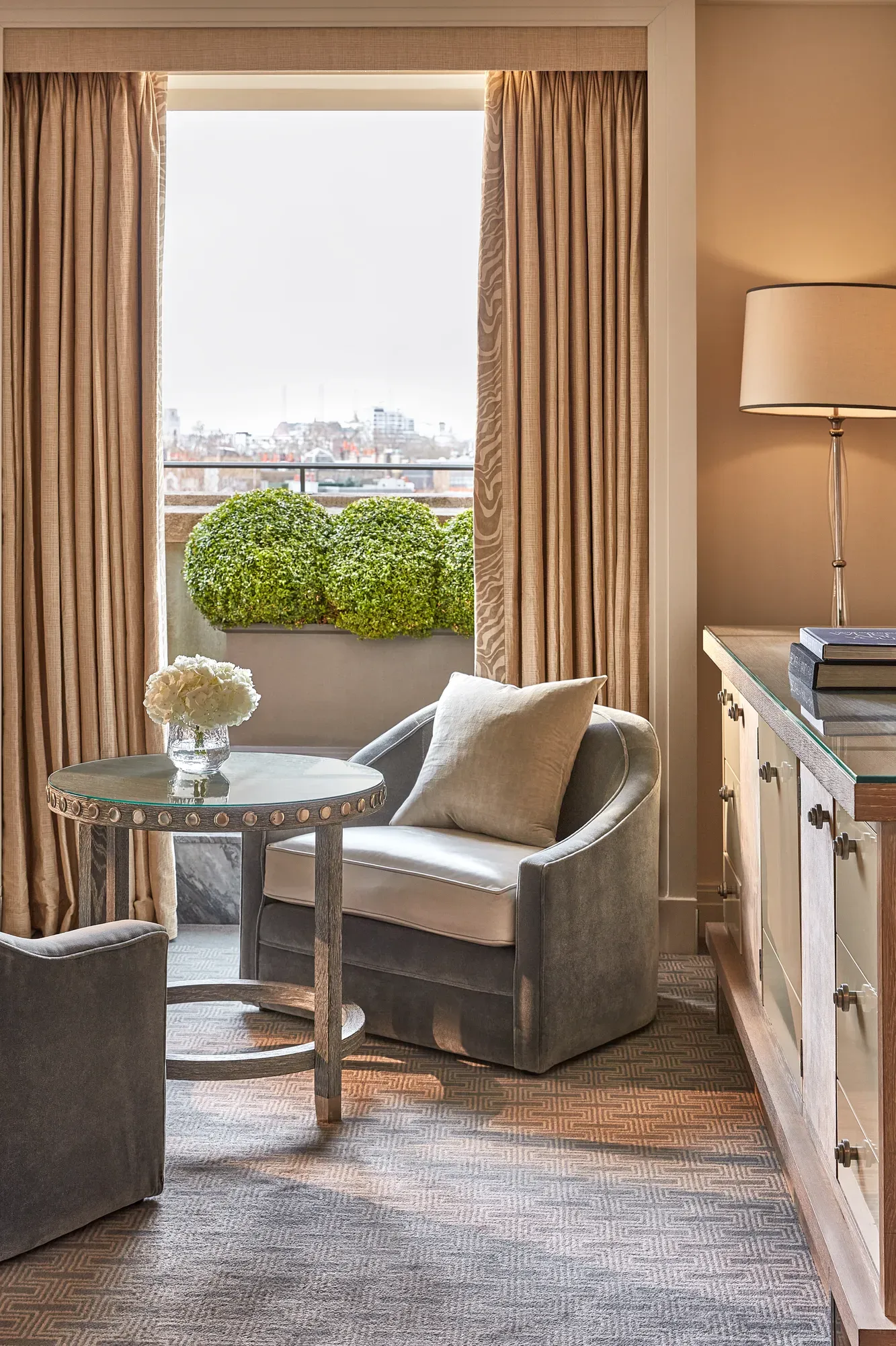 Seating area with two grey armchairs, round table, and balcony view framed by tall curtains.