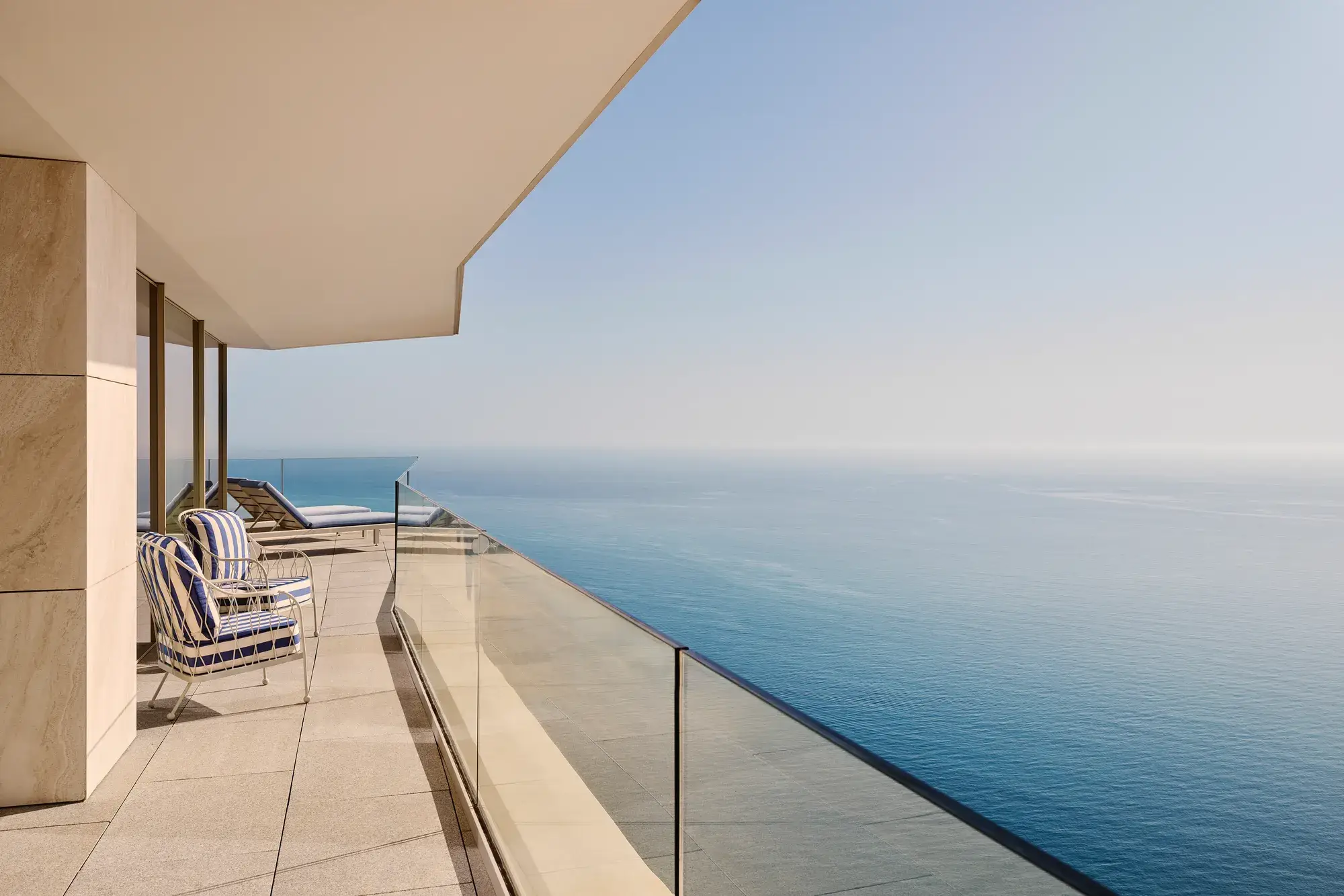 Balcon avec fauteuils rayés et transats derrière un garde-corps en verre, offrant une vue dégagée sur la mer sous un ciel bleu clair.