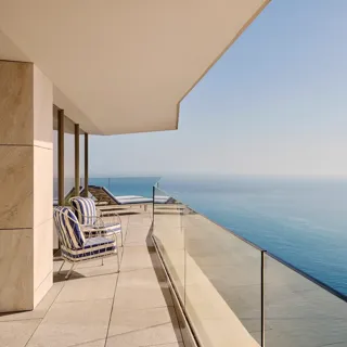 Balcon avec fauteuils rayés et transats derrière un garde-corps en verre, offrant une vue dégagée sur la mer sous un ciel bleu clair.