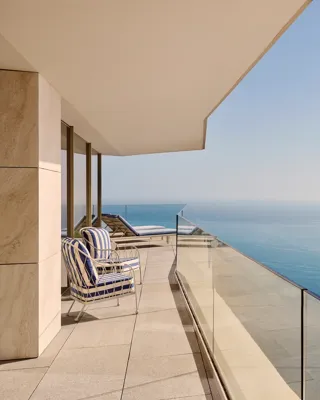 Balcon avec fauteuils rayés et transats derrière un garde-corps en verre, offrant une vue dégagée sur la mer sous un ciel bleu clair.