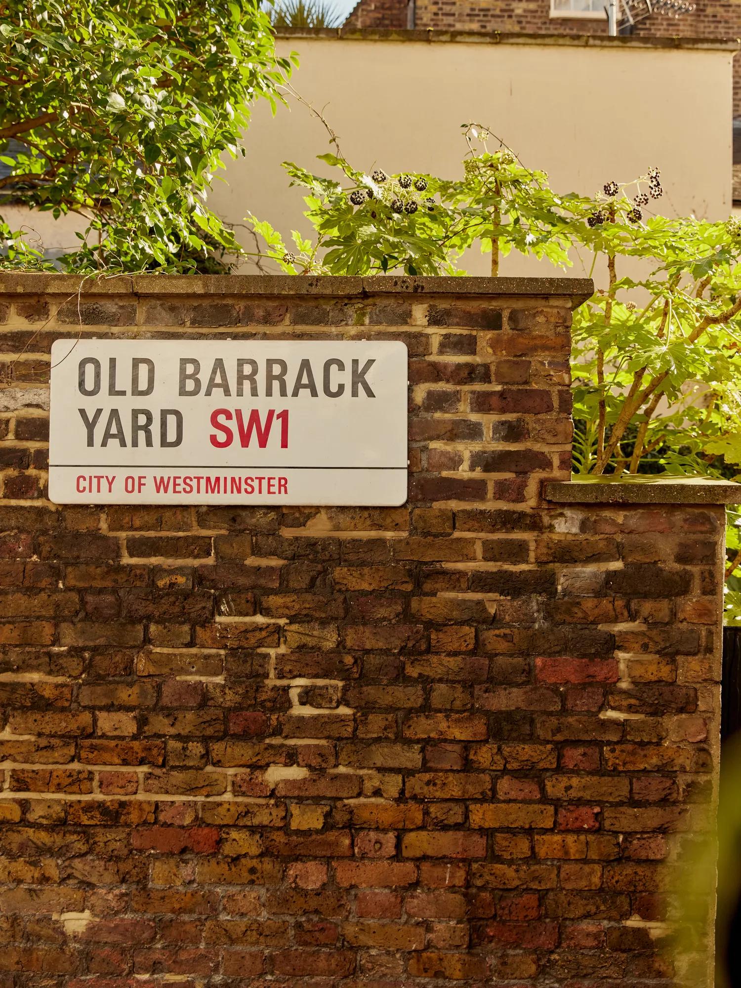 Old Barrack Yard SW1 street sign on a brick wall, surrounded by greenery.