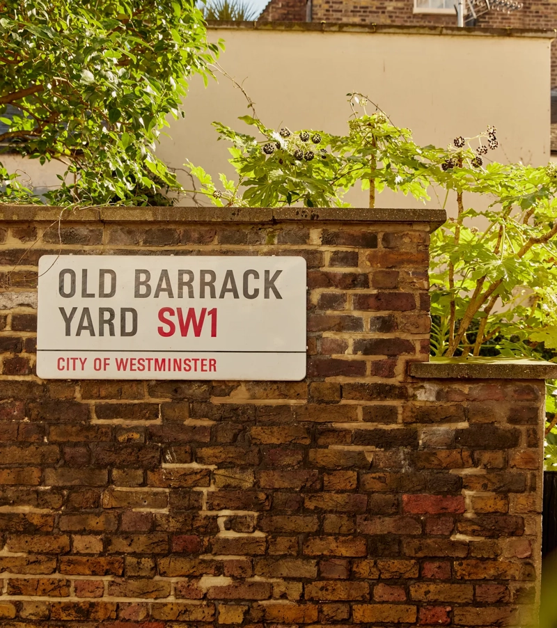 Old Barrack Yard SW1 street sign on a brick wall, surrounded by greenery.