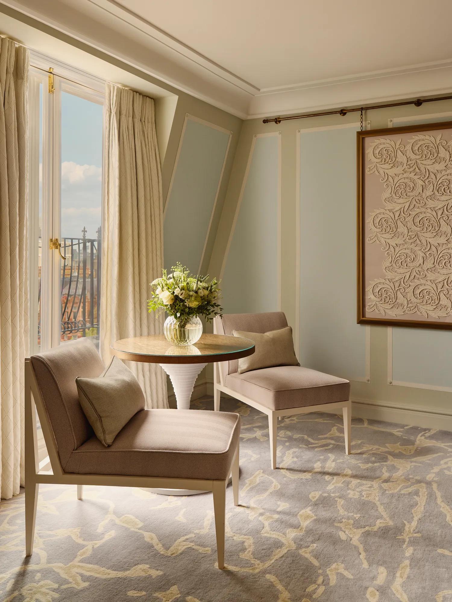 Light-filled sitting area with two upholstered chairs, round table with flowers, patterned carpet, and balcony view through tall windows.