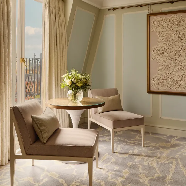 Light-filled sitting area with two upholstered chairs, round table with flowers, patterned carpet, and balcony view through tall windows.