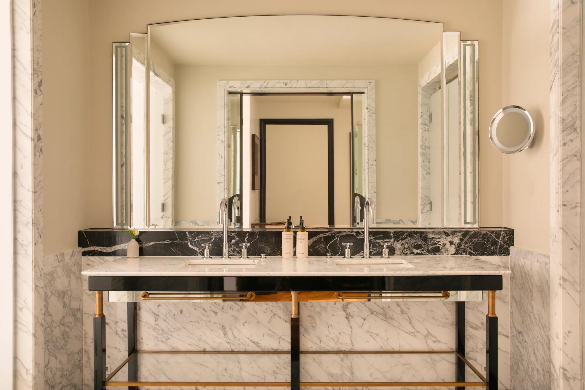 Double marble vanity with illuminated mirrors, twin basins and brass accents in a spacious bathroom.