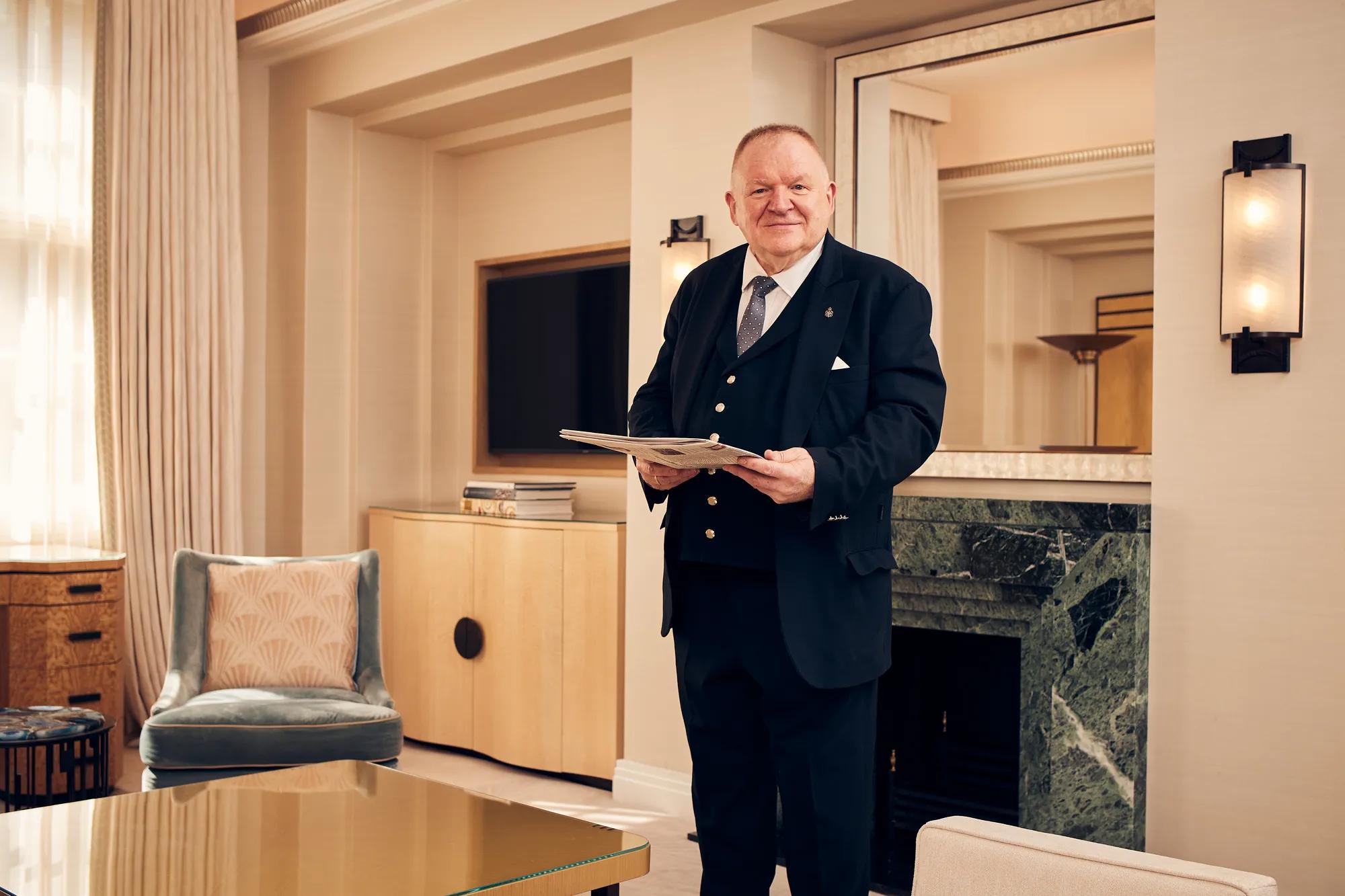 Smiling hotel staff member in formal suit holding a folder in an elegant cream-toned sitting room with a marble fireplace.