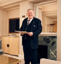 Smiling hotel staff member in formal suit holding a folder in an elegant cream-toned sitting room with a marble fireplace.