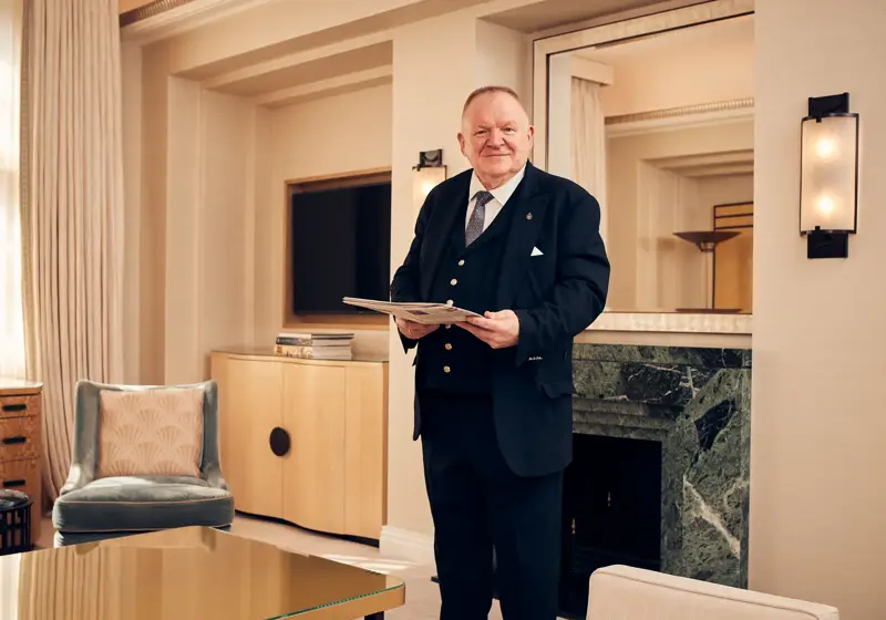 Smiling hotel staff member in formal suit holding a folder in an elegant cream-toned sitting room with a marble fireplace.