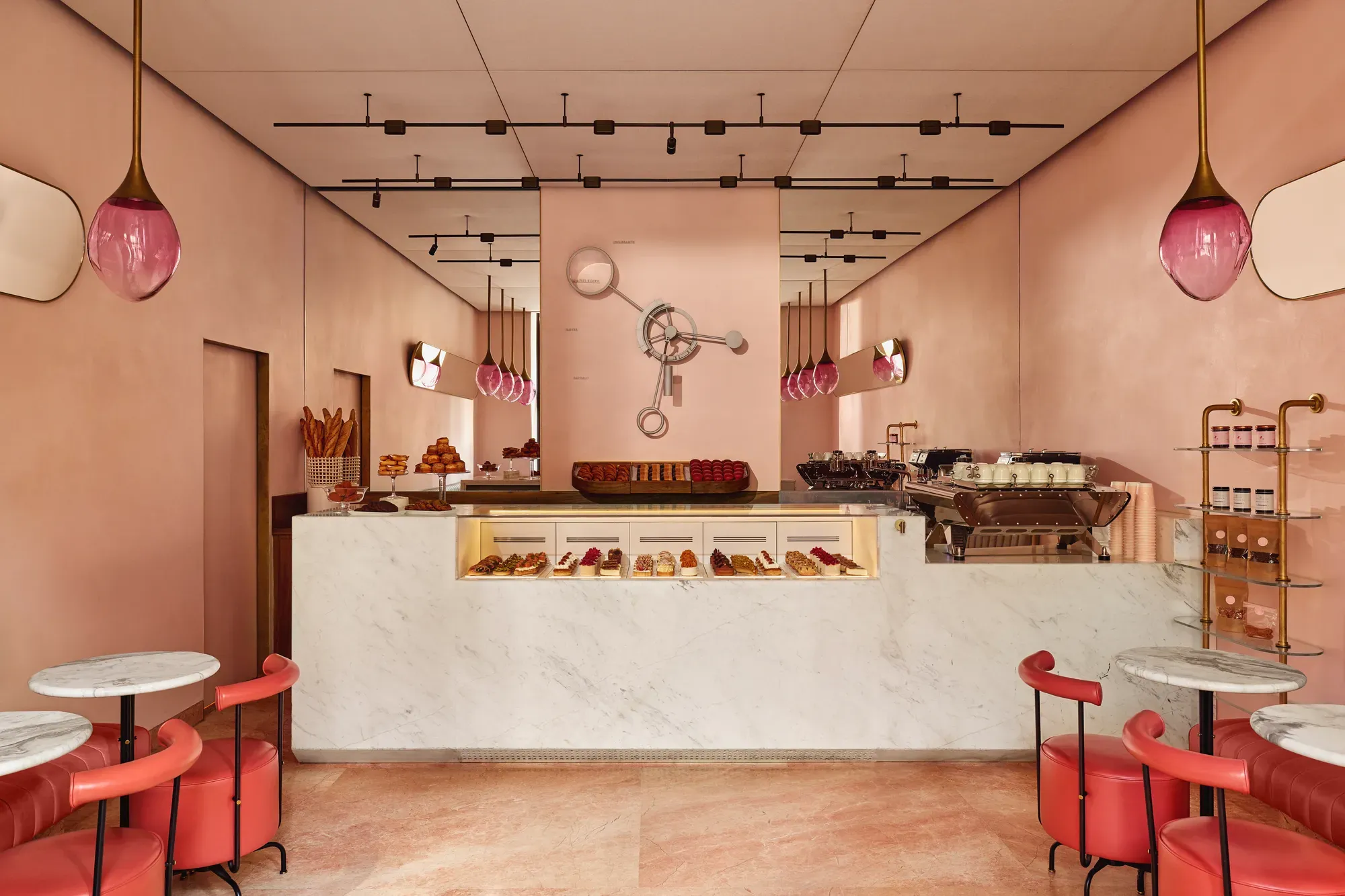 A pink pastry boutique with marble counters, glass pastry displays, red leather seating, and gold light fixtures with pink glass shades.