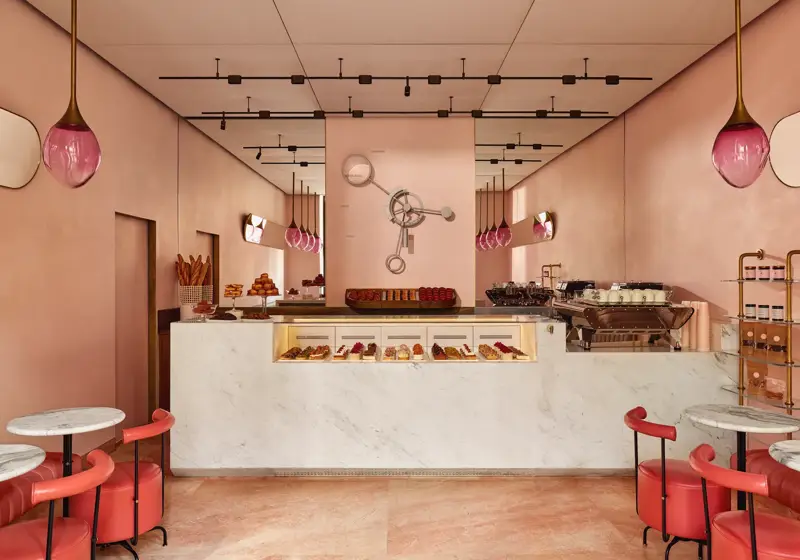 A pink pastry boutique with marble counters, glass pastry displays, red leather seating, and gold light fixtures with pink glass shades.