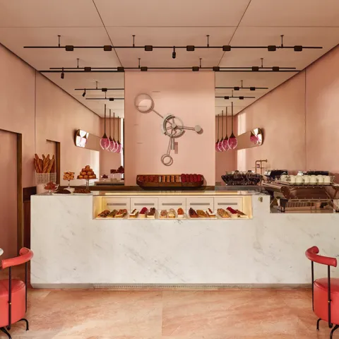 A pink pastry boutique with marble counters, glass pastry displays, red leather seating, and gold light fixtures with pink glass shades.