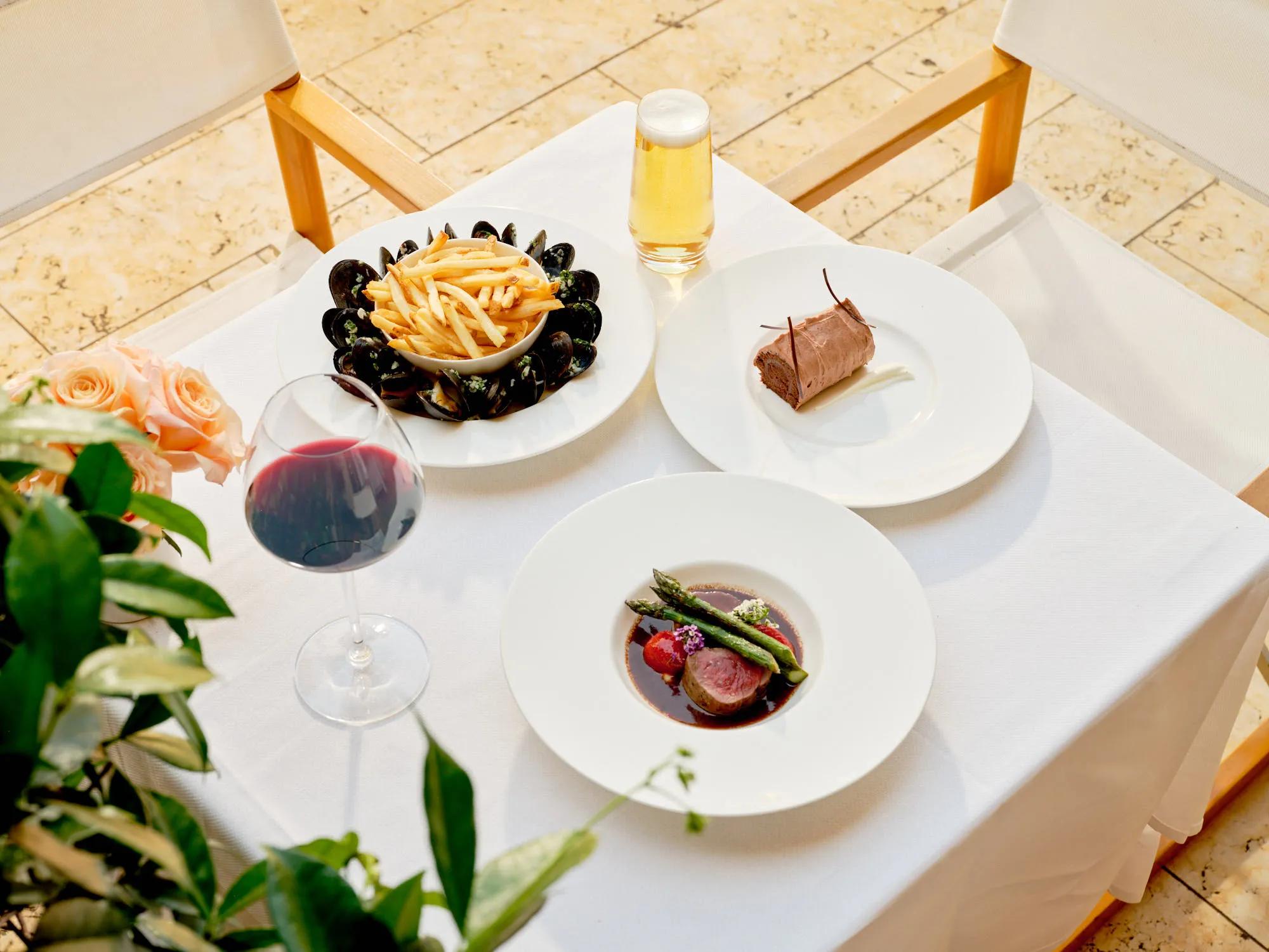 Outdoor table setting with mussels and fries, seared filet with asparagus, dessert, and glasses of red wine and beer.