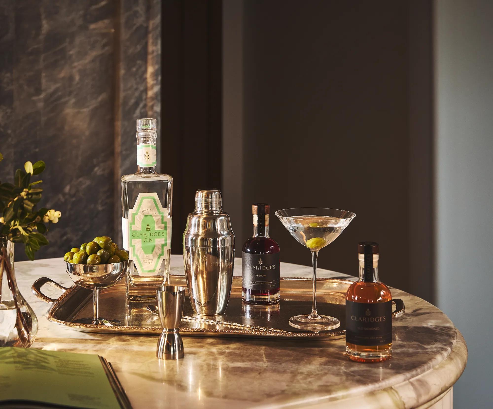 Marble table with cocktail setup, including martini glass, shaker, spirits, olives, and Claridge’s gin bottle.