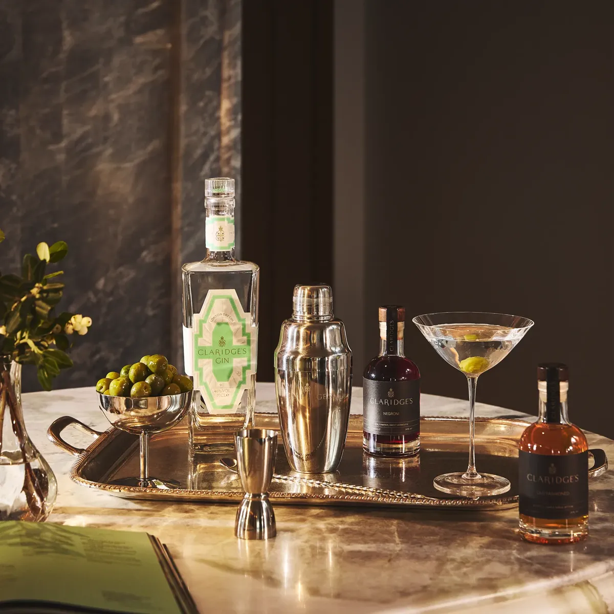 Marble table with cocktail setup, including martini glass, shaker, spirits, olives, and Claridge’s gin bottle.