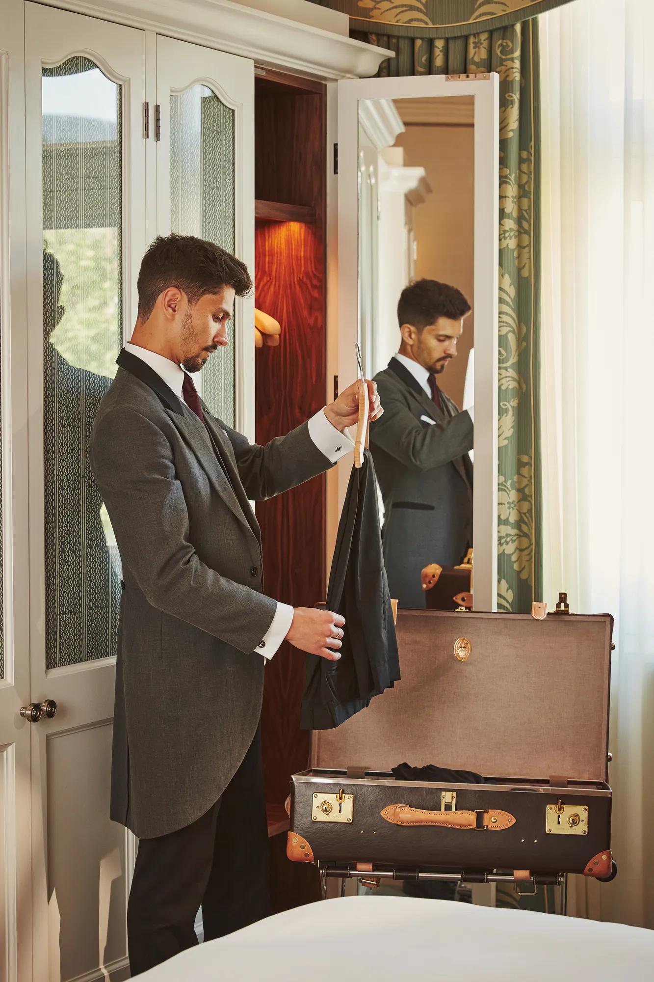 Hotel staff member in a grey tailcoat unpacks a guest’s suitcase, holding a garment hanger beside an open wardrobe.