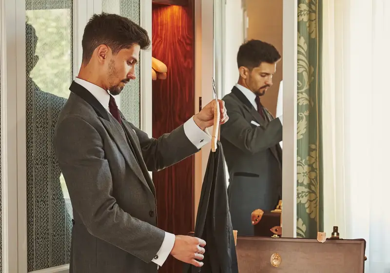 Hotel staff member in a grey tailcoat unpacks a guest’s suitcase, holding a garment hanger beside an open wardrobe.
