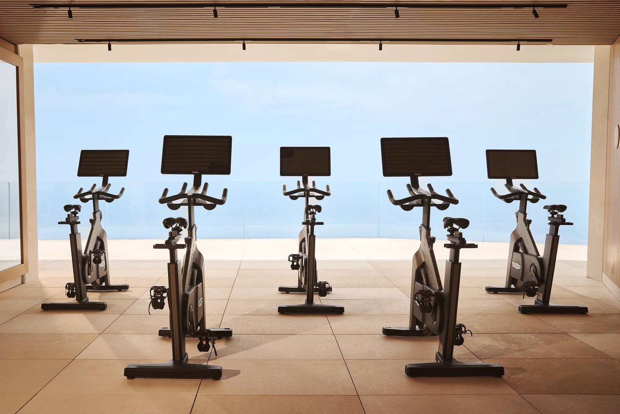 Interior of the Surrenne Riviera open-air fitness studio with a row of indoor exercise bikes positioned on a light-coloured floor, surrounded by natural elements, creating a spacious and airy atmosphere.