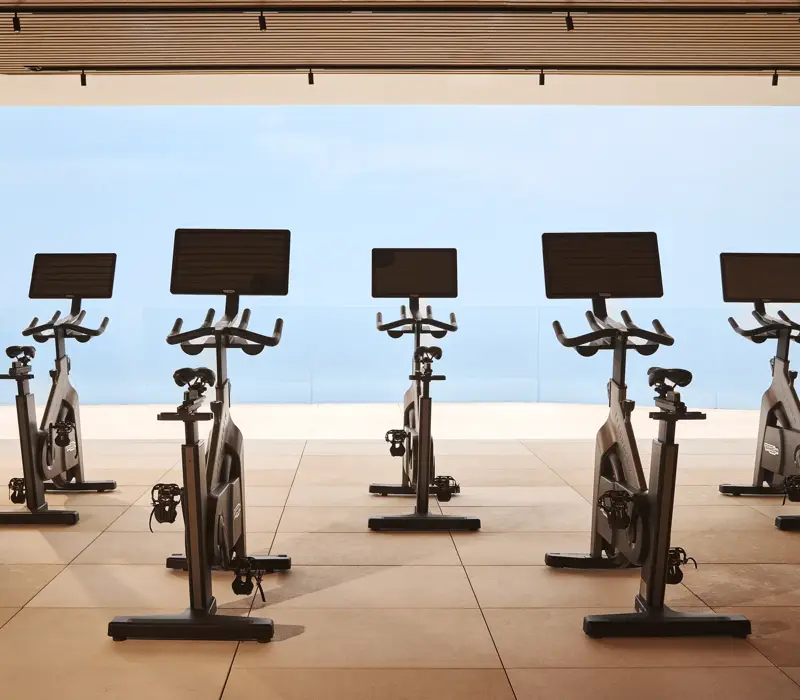 Interior of the Surrenne Riviera open-air fitness studio with a row of indoor exercise bikes positioned on a light-coloured floor, surrounded by natural elements, creating a spacious and airy atmosphere.