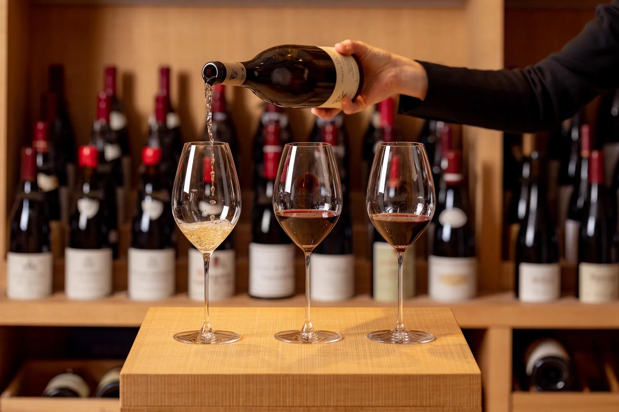 Hand pouring white wine into a glass beside two filled red wine glasses, with wine bottles in the background.