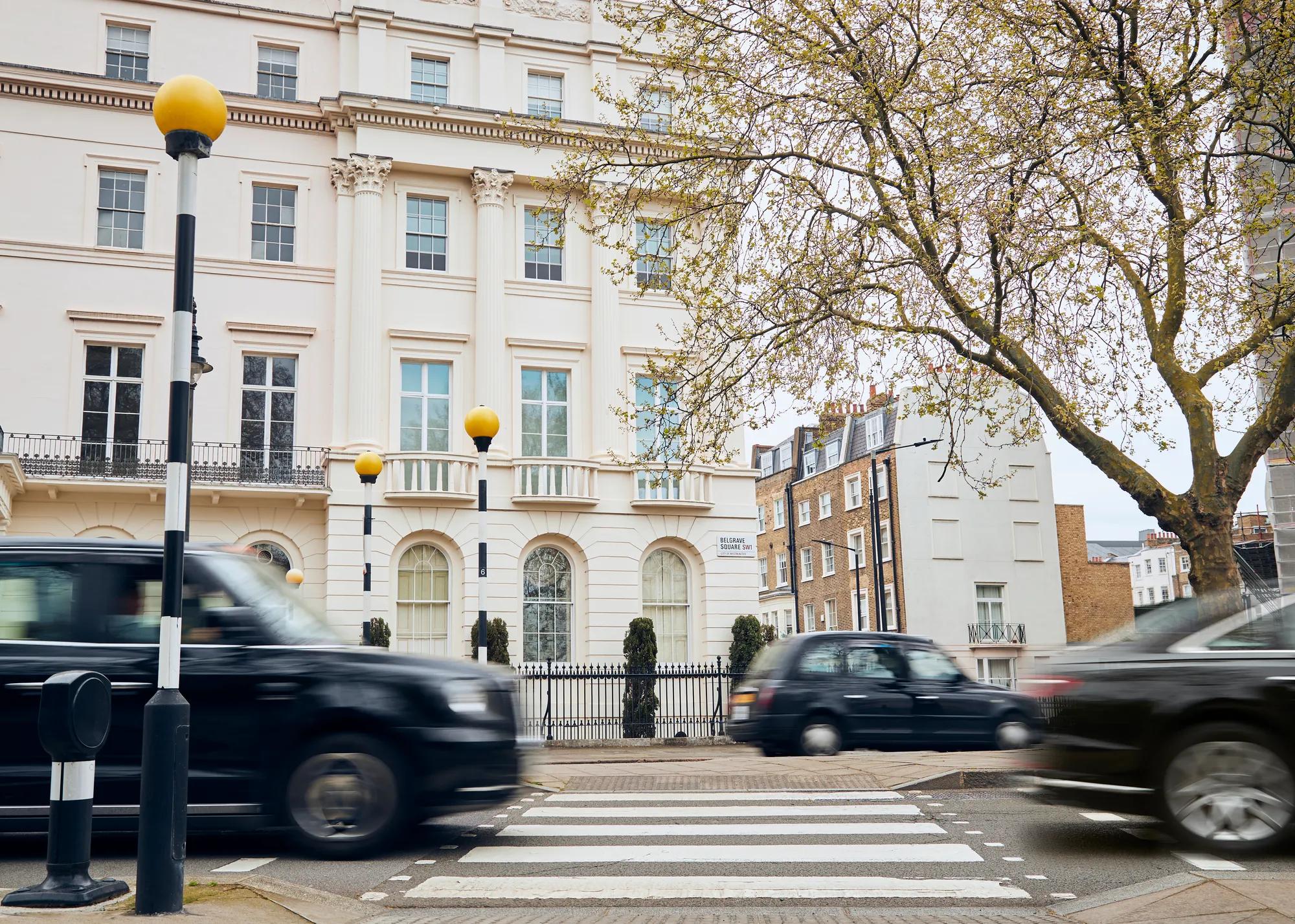 Black cabs and cars drive past a pedestrian crossing in front of a grand white townhouse on a tree-lined London street.