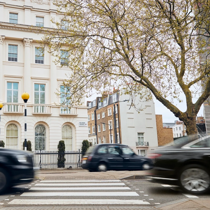 Black cabs and cars drive past a pedestrian crossing in front of a grand white townhouse on a tree-lined London street.
