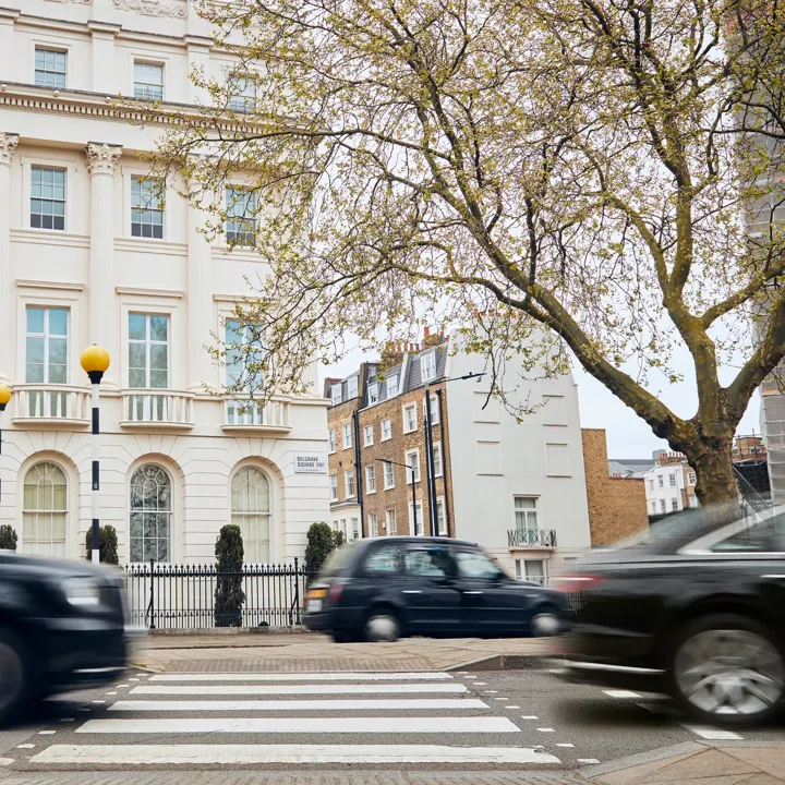 Black cabs and cars drive past a pedestrian crossing in front of a grand white townhouse on a tree-lined London street.
