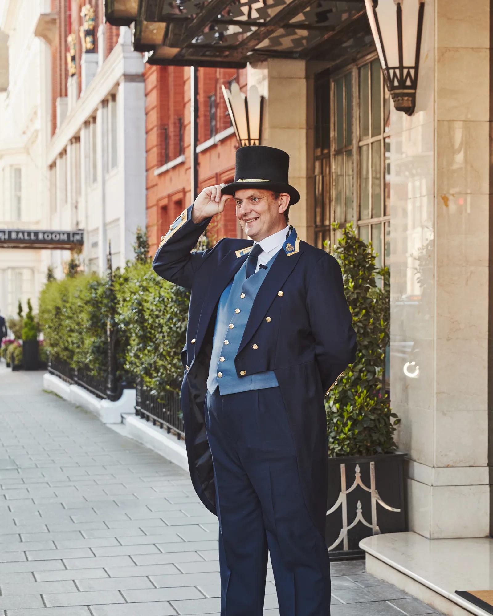 A Claridge’s doorman in a navy uniform and top hat smiling and tipping his hat outside the hotel entrance on a sunny London street.
