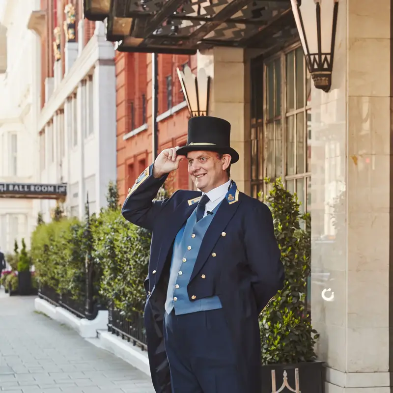 A Claridge’s doorman in a navy uniform and top hat smiling and tipping his hat outside the hotel entrance on a sunny London street.