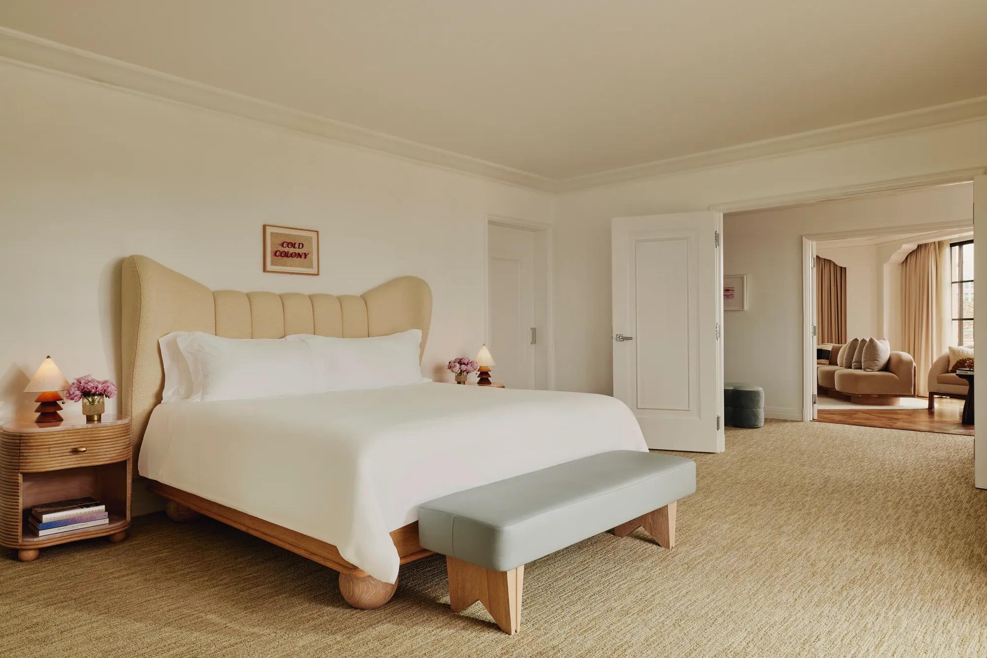Primary bedroom with winged upholstered headboard, pale leather bench, wood nightstands with sculptural lamps and pink roses; double doors open to living area.