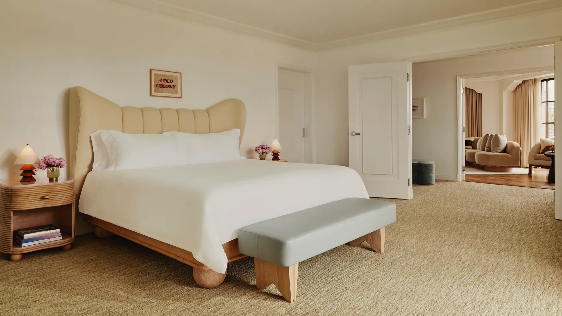 Primary bedroom with winged upholstered headboard, pale leather bench, wood nightstands with sculptural lamps and pink roses; double doors open to living area.