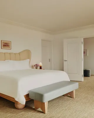 Primary bedroom with winged upholstered headboard, pale leather bench, wood nightstands with sculptural lamps and pink roses; double doors open to living area.