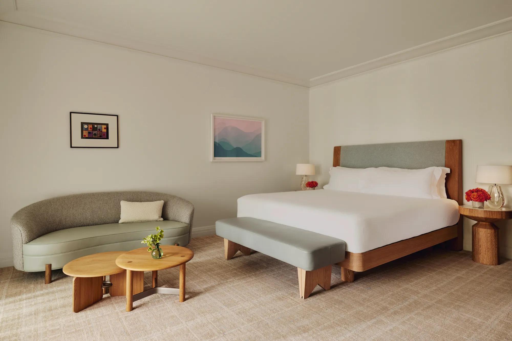 Guestroom with king bed and bench, adjacent seating area with curved sofa and nested tables, framed art, and floor-to-ceiling curtains.