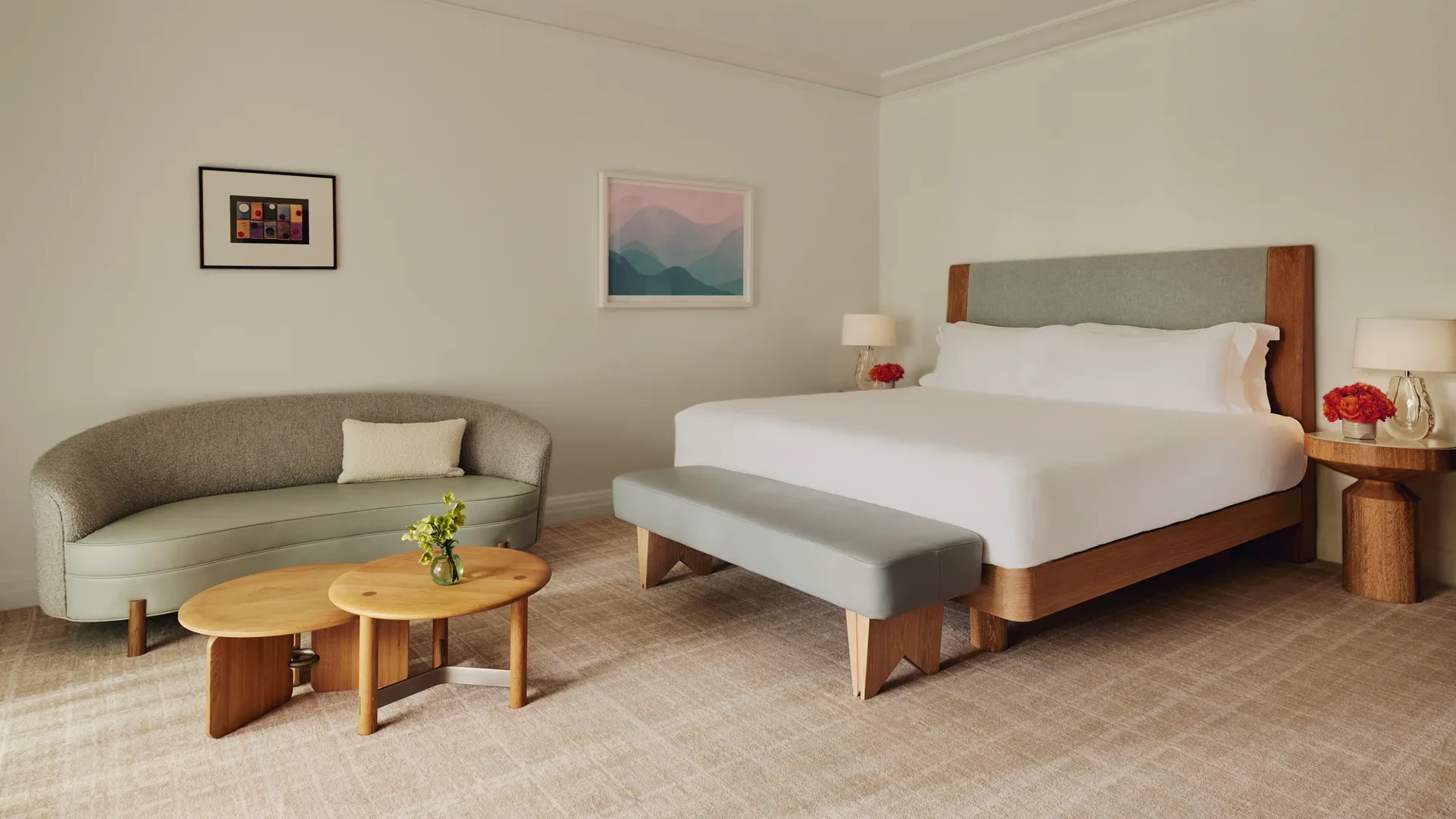 Guestroom with king bed and bench, adjacent seating area with curved sofa and nested tables, framed art, and floor-to-ceiling curtains.