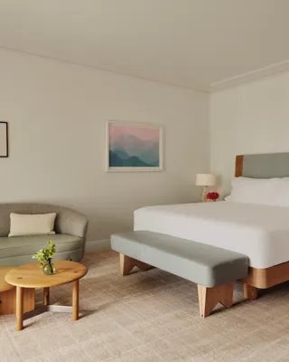 Guestroom with king bed and bench, adjacent seating area with curved sofa and nested tables, framed art, and floor-to-ceiling curtains.