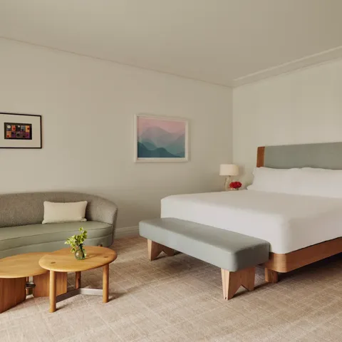 Guestroom with king bed and bench, adjacent seating area with curved sofa and nested tables, framed art, and floor-to-ceiling curtains.