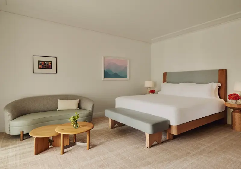 Guestroom with king bed and bench, adjacent seating area with curved sofa and nested tables, framed art, and floor-to-ceiling curtains.