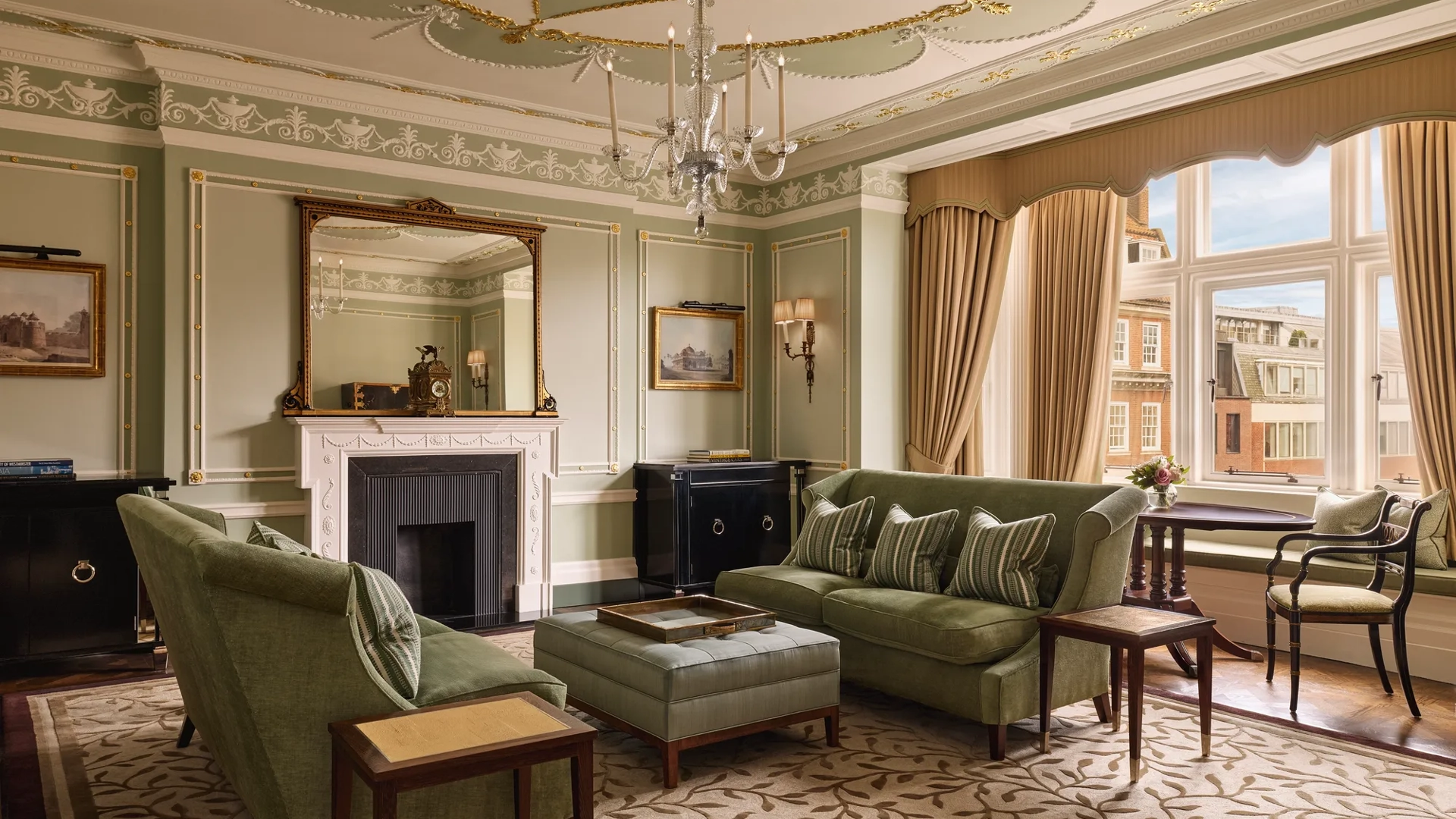 An elegant sitting room with sage green walls, gold detailing, and classic furnishings including velvet sofas, a marble fireplace, chandelier, and large window overlooking red-brick Mayfair buildings.