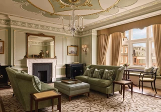 An elegant sitting room with sage green walls, gold detailing, and classic furnishings including velvet sofas, a marble fireplace, chandelier, and large window overlooking red-brick Mayfair buildings.
