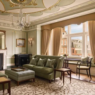An elegant sitting room with sage green walls, gold detailing, and classic furnishings including velvet sofas, a marble fireplace, chandelier, and large window overlooking red-brick Mayfair buildings.