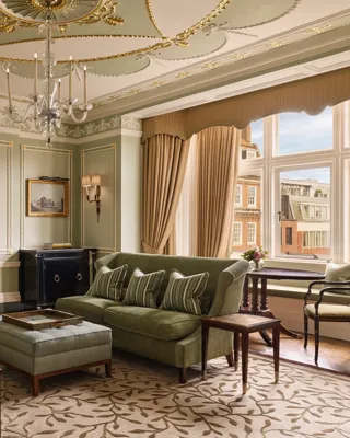 An elegant sitting room with sage green walls, gold detailing, and classic furnishings including velvet sofas, a marble fireplace, chandelier, and large window overlooking red-brick Mayfair buildings.